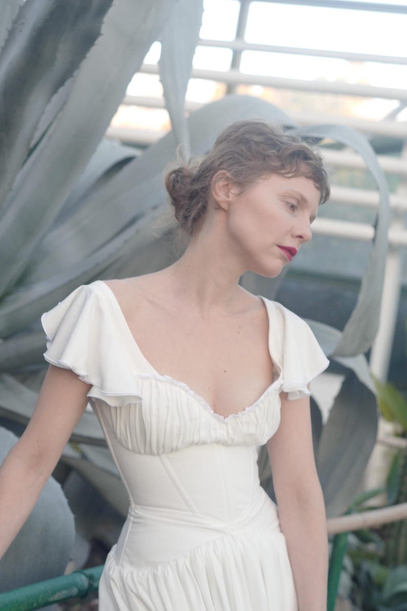 May include: A white dress with a fitted bodice and a flowing skirt. The dress features ruffled sleeves and a low neckline. The woman in the image has red lipstick and her hair is styled in a bun. The background includes large green plants.