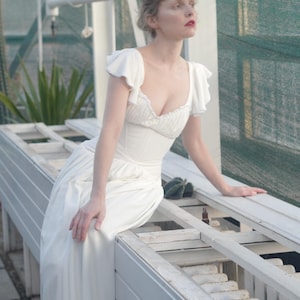 May include: A white dress with a fitted bodice and flowing skirt. The dress features short, ruffled sleeves and a low neckline. The person is sitting on a white bench in a greenhouse setting, with a green mesh background and a few plants.