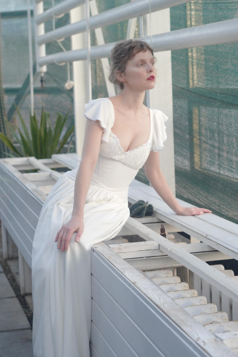 May include: A white dress with a fitted bodice and flowing skirt. The dress features short, ruffled sleeves and a low neckline. The person is sitting on a white bench in a greenhouse setting, with a green mesh background and a few plants.