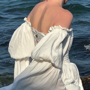 Puede incluir: Un vestido blanco de hombros descubiertos con mangas abullonadas y un detalle de encaje en la espalda. El vestido fluye con el viento, creando un look rom&aacute;ntico y et&eacute;reo.