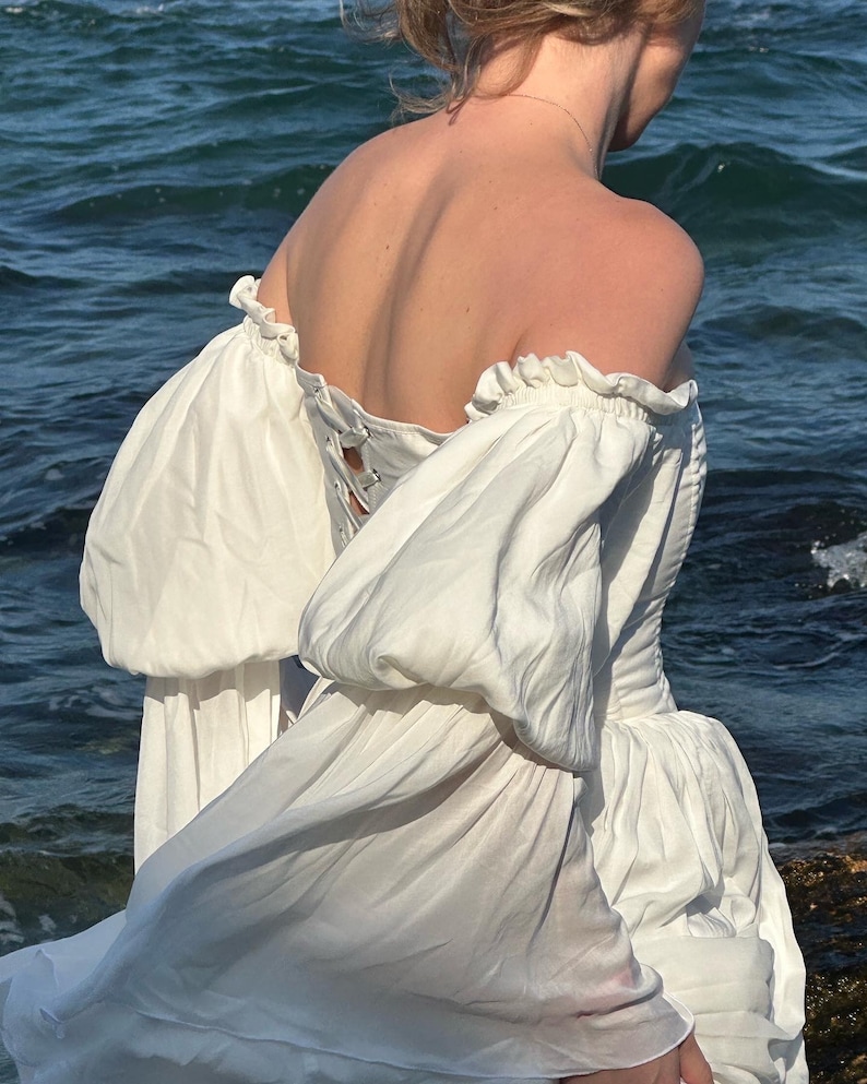 Puede incluir: Un vestido blanco de hombros descubiertos con mangas abullonadas y un detalle de encaje en la espalda. El vestido fluye con el viento, creando un look rom&aacute;ntico y et&eacute;reo.