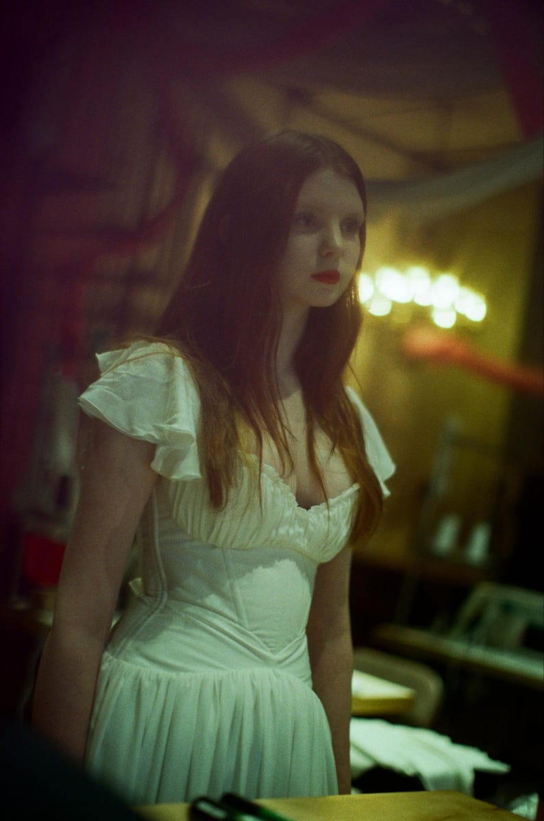 May include: A woman in a white dress with a fitted bodice and flowing skirt. The dress has ruffled sleeves and a low neckline. The woman has long, reddish-brown hair and is wearing red lipstick. The background is dimly lit.