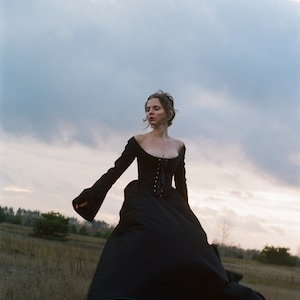 May include: A floor-length black gown featuring a fitted bodice with a lace-up front closure, a wide scoop neckline, and long, flared sleeves. The skirt is voluminous and full, extending to the ground with a structured silhouette.