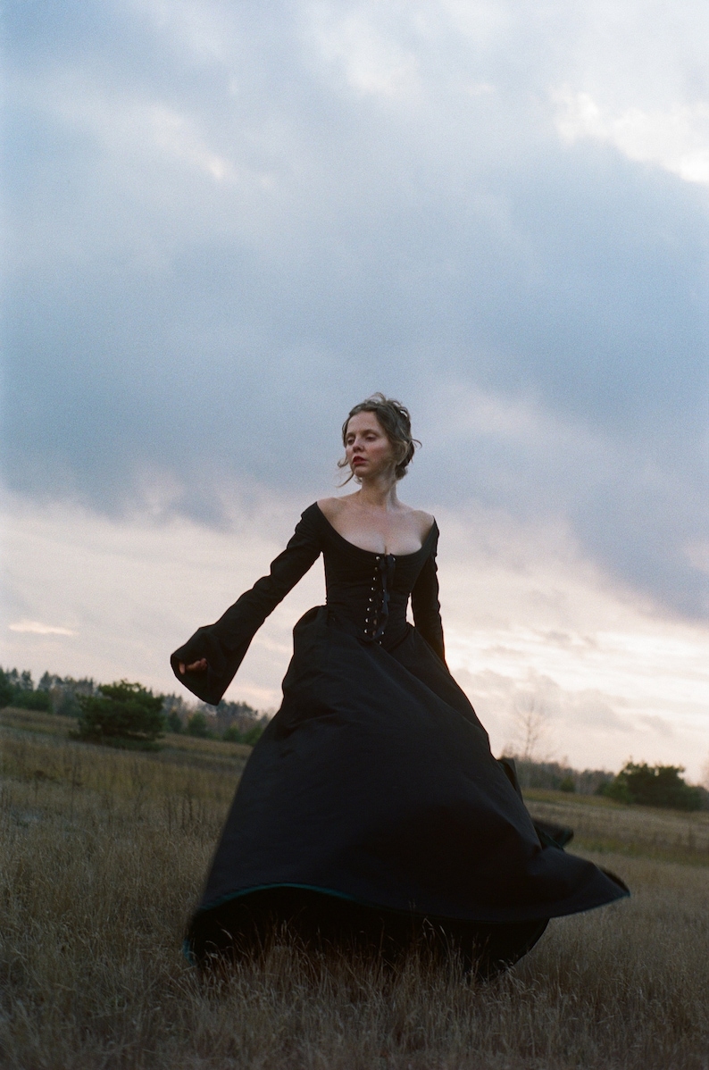 May include: A floor-length black gown featuring a fitted bodice with a lace-up front closure, a wide scoop neckline, and long, flared sleeves. The skirt is voluminous and full, extending to the ground with a structured silhouette.