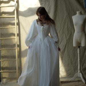 May include: A white, off-the-shoulder dress with long, puffy sleeves and a corset-style bodice. The dress has a flowing skirt and is made of a sheer, lightweight fabric. A wooden ladder and dress form are in the background.