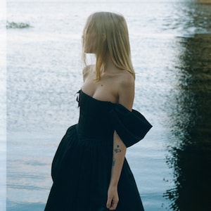 May include: A woman wearing a black off-the-shoulder dress with a fitted bodice and a full skirt. She is standing by a body of water.