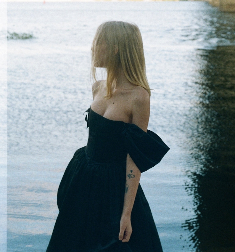 May include: A woman wearing a black off-the-shoulder dress with a fitted bodice and a full skirt. She is standing by a body of water.
