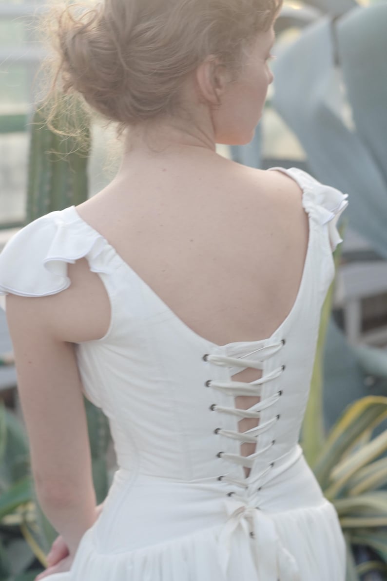 May include: A white dress with a corset back and ruffled sleeves. The dress is made of a soft fabric and has a low back. The back of the dress is laced up with a white ribbon. The dress is a vintage style.