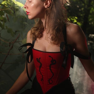 May include: A woman wearing a red corset with black ribbon straps and a black skirt. The corset has a snake design on the front. The woman is standing in a forest setting with smoke in the background.