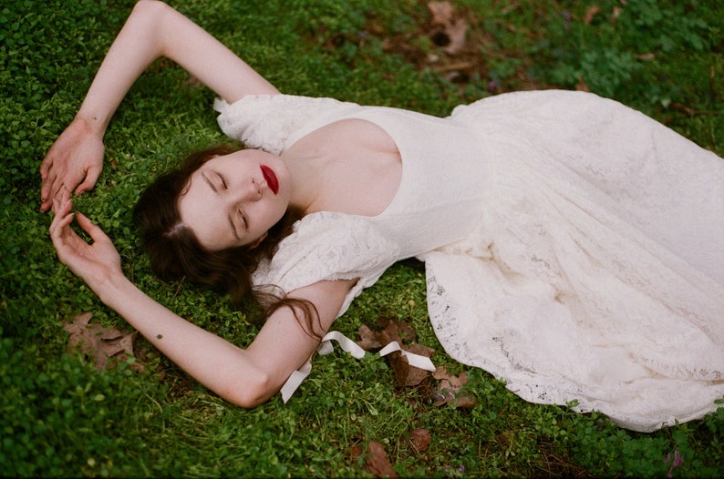 May include: A woman wearing a white lace dress with a low neckline, lying on her back on a bed of green grass.