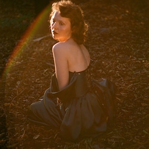 May include: A woman wearing a dark gray strapless dress sits on a bed of brown leaves. The sun shines behind her, creating a rainbow effect.