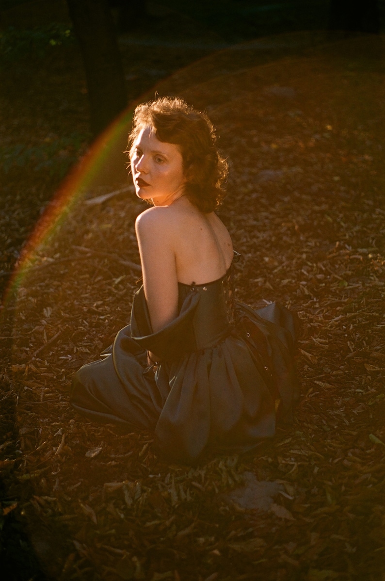 May include: A woman wearing a dark gray strapless dress sits on a bed of brown leaves. The sun shines behind her, creating a rainbow effect.