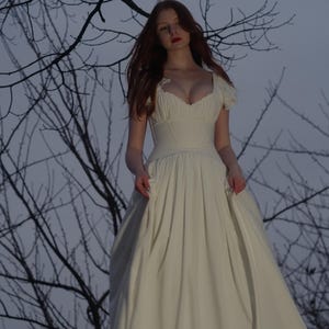 May include: A long, off-white dress with a fitted bodice and a flowing skirt. The dress features a sweetheart neckline and short, puffed sleeves. The background includes bare tree branches against a dusky sky, creating a romantic and ethereal aesthetic.