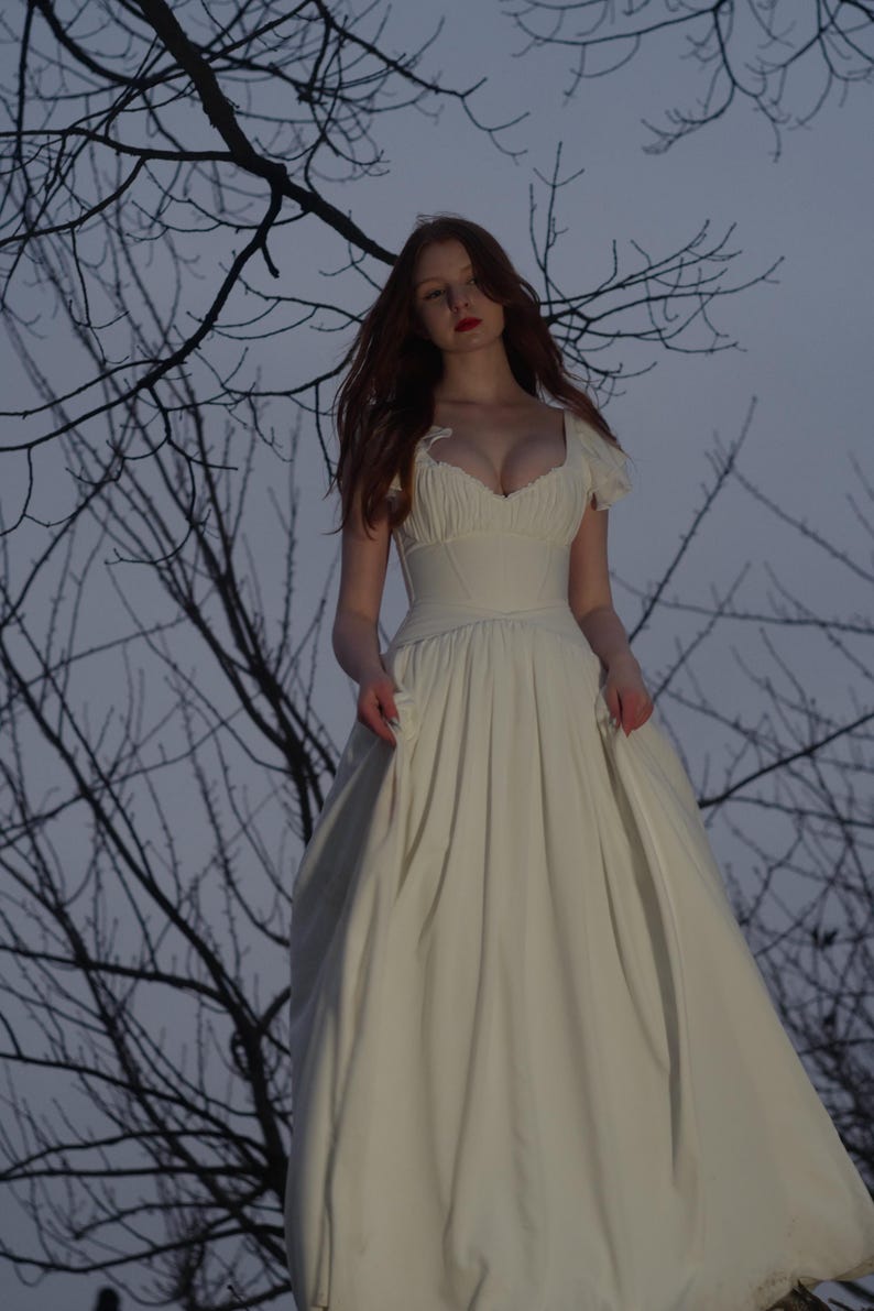 May include: A long, off-white dress with a fitted bodice and a flowing skirt. The dress features a sweetheart neckline and short, puffed sleeves. The background includes bare tree branches against a dusky sky, creating a romantic and ethereal aesthetic.