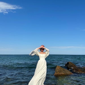 Puede incluir: Una mujer con un vestido blanco maxi de manga larga se encuentra de pie sobre una formaci&oacute;n rocosa junto al mar. El cielo es azul y el agua es de un azul profundo.