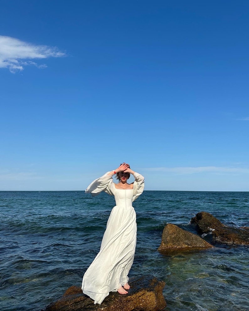 Puede incluir: Una mujer con un vestido blanco maxi de manga larga se encuentra de pie sobre una formaci&oacute;n rocosa junto al mar. El cielo es azul y el agua es de un azul profundo.