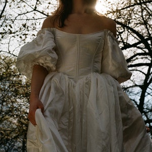 Puede incluir: Una mujer con un vestido blanco de hombros descubiertos con mangas abullonadas y un corpi&ntilde;o ajustado. El vestido es fluido y se agita con el viento. La mujer est&aacute; de pie en un bosque con &aacute;rboles en el fondo.