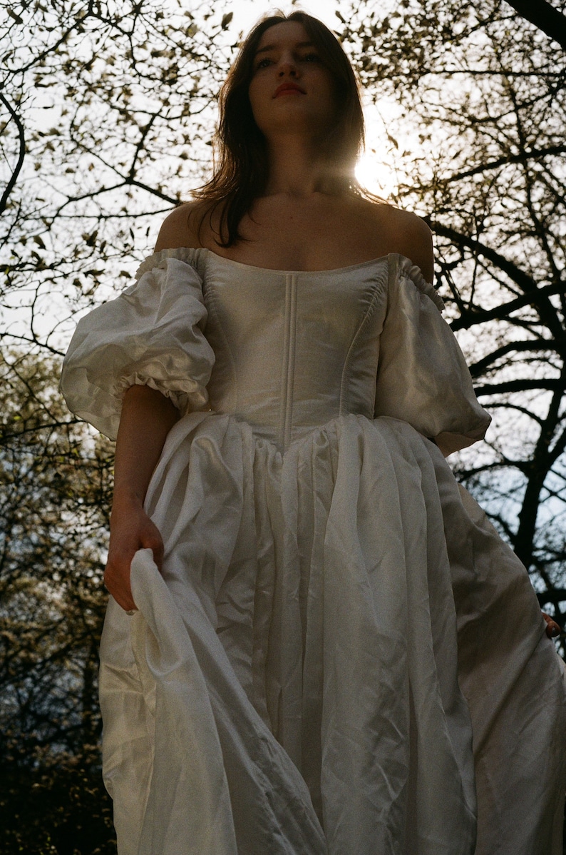 Puede incluir: Una mujer con un vestido blanco de hombros descubiertos con mangas abullonadas y un corpi&ntilde;o ajustado. El vestido es fluido y se agita con el viento. La mujer est&aacute; de pie en un bosque con &aacute;rboles en el fondo.