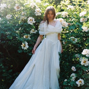 May include: A woman's white dress with a sweetheart neckline and flowing skirt. The dress is worn in a garden with white flowers and green foliage. The dress has a vintage or romantic style.