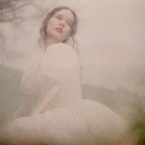May include: A woman wearing a white dress with a white puff sleeve. The woman is standing in a field with a blurry background.