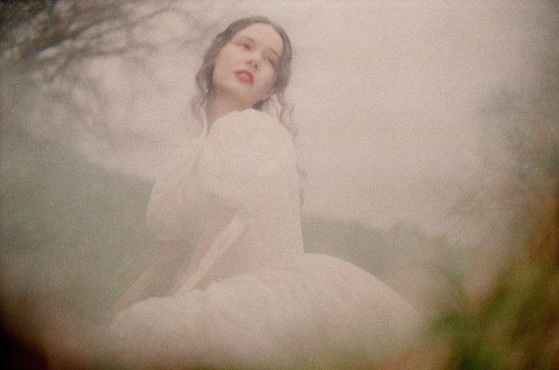 May include: A woman wearing a white dress with a white puff sleeve. The woman is standing in a field with a blurry background.