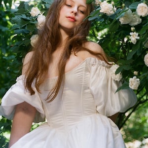 Puede incluir: Una mujer con un vestido blanco de hombros descubiertos con mangas abullonadas se encuentra en un jard&iacute;n con flores blancas.