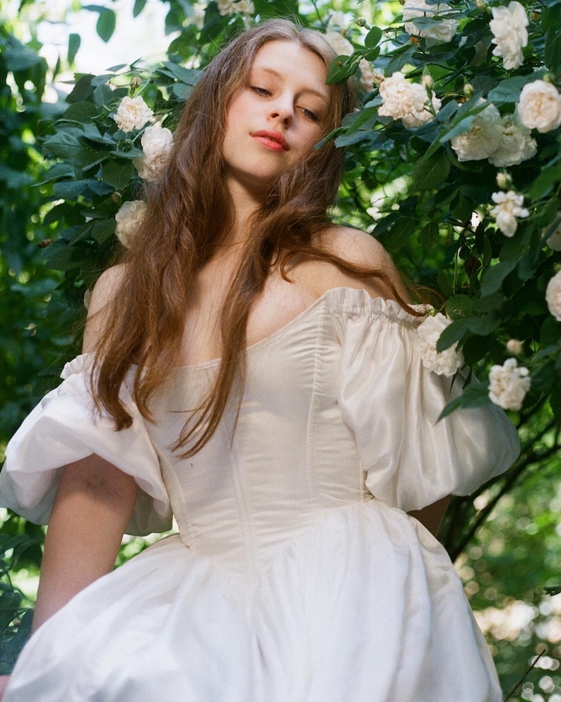Puede incluir: Una mujer con un vestido blanco de hombros descubiertos con mangas abullonadas se encuentra en un jard&iacute;n con flores blancas.