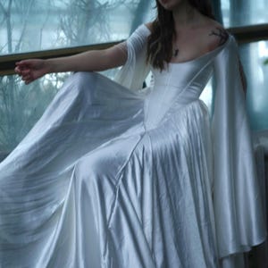 Könnte beinhalten: Weißes Satinkleid mit eng anliegendem Oberteil und fließendem Rock. Das Kleid hat schulterfreie Ärmel und eine lange, elegante Silhouette. Das Model sitzt in der Nähe eines Fensters und erzeugt eine weiche, ätherische Ästhetik.