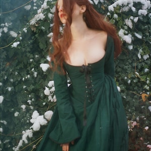 May include: A long-sleeved, floor-length dark green dress featuring a wide scoop neckline and a central black lace-up corset-style bodice with a ribbon tie. The garment has fitted sleeves and a full, flowing skirt with a structured, fitted waistline.
