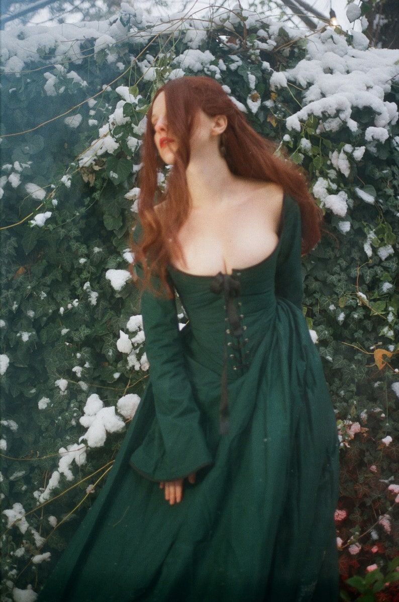 May include: A long-sleeved, floor-length dark green dress featuring a wide scoop neckline and a central black lace-up corset-style bodice with a ribbon tie. The garment has fitted sleeves and a full, flowing skirt with a structured, fitted waistline.