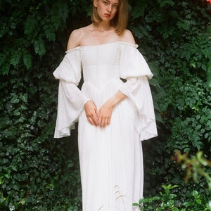 Puede incluir: Una mujer con un vestido blanco de hombros descubiertos con mangas abullonadas, de pie frente a un fondo verde frondoso.