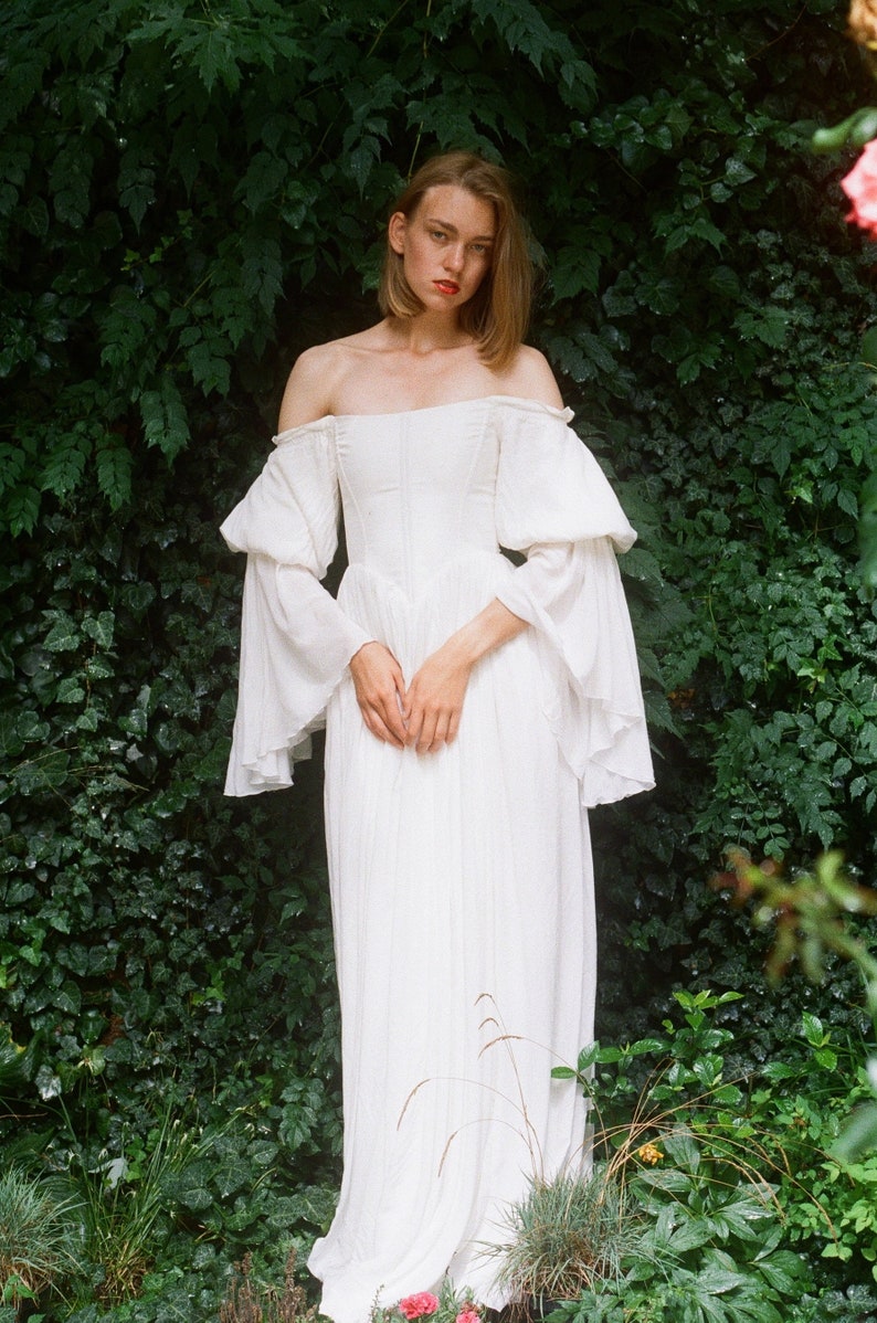 Puede incluir: Una mujer con un vestido blanco de hombros descubiertos con mangas abullonadas, de pie frente a un fondo verde frondoso.
