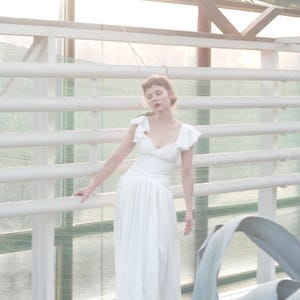 May include: A white, floor-length dress with a fitted bodice and flowing skirt. The dress features ruffled cap sleeves and a V-neckline. The image is set in a greenhouse with white railings and natural light.