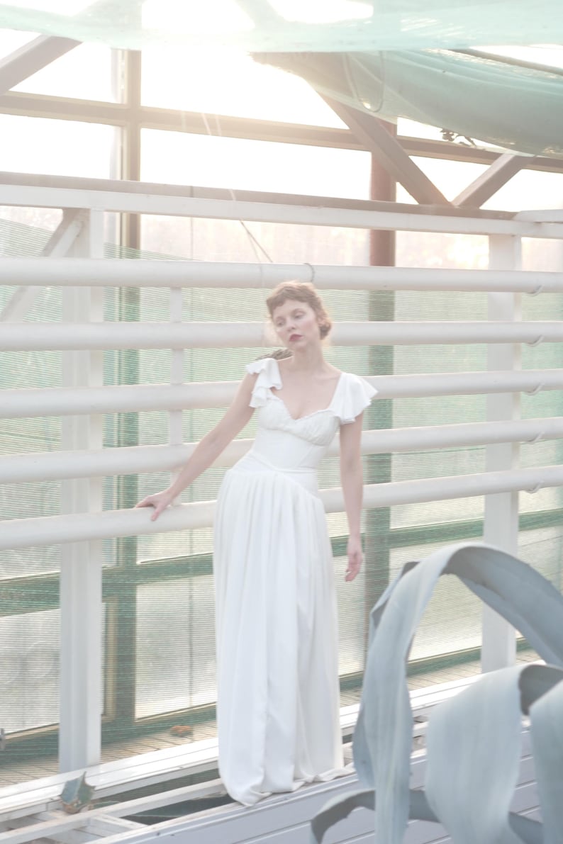 May include: A white, floor-length dress with a fitted bodice and flowing skirt. The dress features ruffled cap sleeves and a V-neckline. The image is set in a greenhouse with white railings and natural light.
