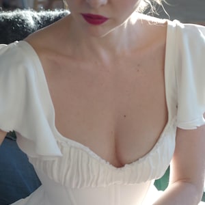 May include: A white dress with a sweetheart neckline and ruffled sleeves. The bodice is fitted, and the dress appears to be made of a soft, flowing fabric. The model has red lipstick.