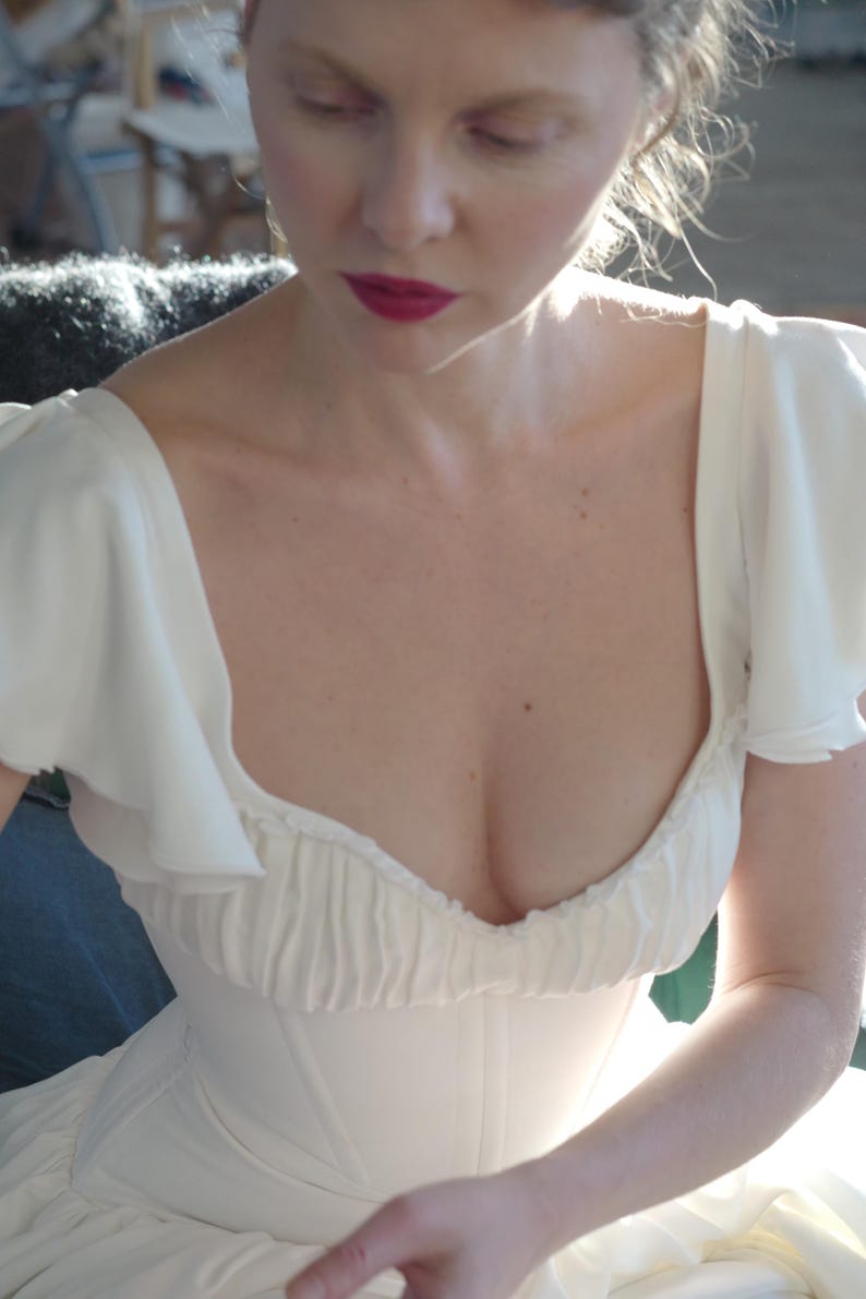 May include: A white dress with a sweetheart neckline and ruffled sleeves. The bodice is fitted, and the dress appears to be made of a soft, flowing fabric. The model has red lipstick.
