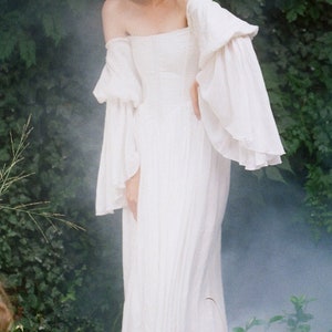 Puede incluir: Una mujer con un vestido blanco sin tirantes y mangas largas y fluidas se encuentra en un jard&iacute;n. El vestido tiene un dise&ntilde;o simple y elegante y est&aacute; hecho de una tela ligera. El cabello de la mujer est&aacute; recogido hacia atr&aacute;s de su rostro, y tiene un aspecto suave y natural.