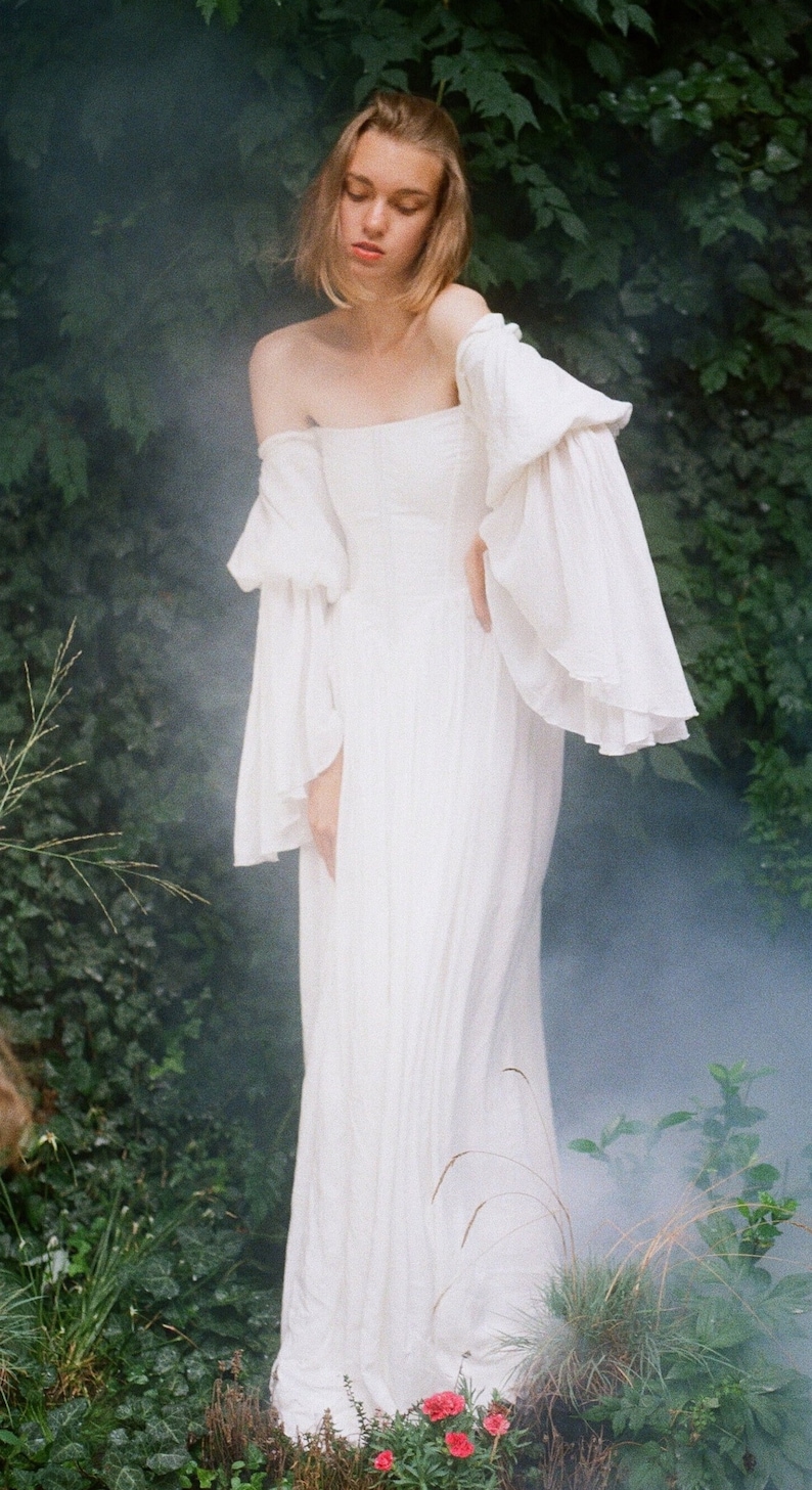 Puede incluir: Una mujer con un vestido blanco sin tirantes y mangas largas y fluidas se encuentra en un jard&iacute;n. El vestido tiene un dise&ntilde;o simple y elegante y est&aacute; hecho de una tela ligera. El cabello de la mujer est&aacute; recogido hacia atr&aacute;s de su rostro, y tiene un aspecto suave y natural.