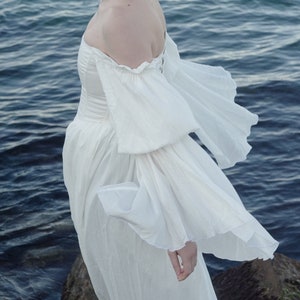 Puede incluir: Una mujer con un vestido blanco de hombros descubiertos y mangas abullonadas, de pie junto al borde del agua.