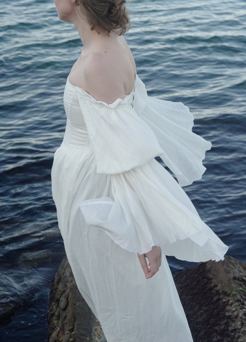 Puede incluir: Una mujer con un vestido blanco de hombros descubiertos y mangas abullonadas, de pie junto al borde del agua.