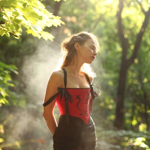 May include: A woman in a red corset with black trim and a black skirt stands in a forest. The corset has a black and red floral design. The woman's hair is pulled back and she is looking down.