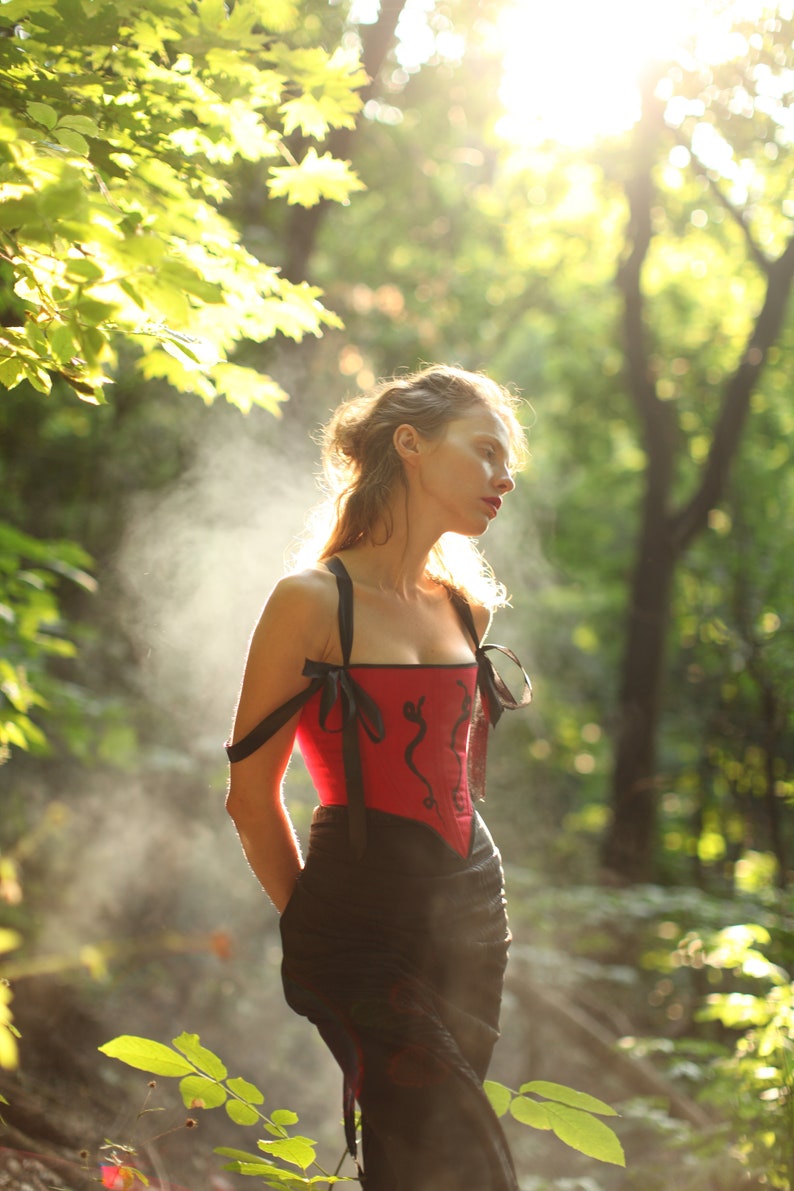 May include: A woman in a red corset with black trim and a black skirt stands in a forest. The corset has a black and red floral design. The woman's hair is pulled back and she is looking down.