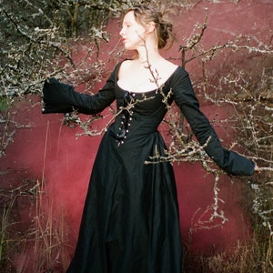 May include: A floor-length black dress featuring long, flared sleeves, a wide scoop neckline, and a central front bodice panel with a vertical row of silver-toned eyelets and a black ribbon lace-up closure that extends to the waistline.
