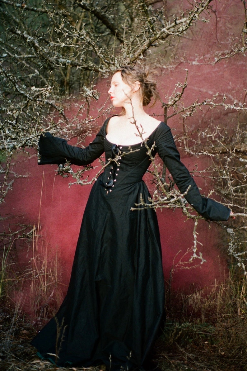 May include: A floor-length black dress featuring long, flared sleeves, a wide scoop neckline, and a central front bodice panel with a vertical row of silver-toned eyelets and a black ribbon lace-up closure that extends to the waistline.