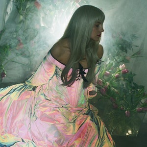 May include: An iridescent, off-the-shoulder gown with a corset-style bodice featuring black lace-up detailing. The fabric displays a shifting, holographic sheen of pink, gold, and blue tones, paired with long, voluminous sleeves and a full skirt.