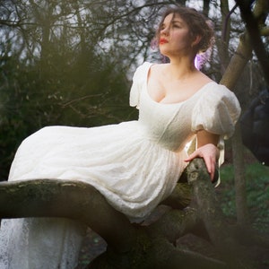May include: A woman wearing a white lace dress with puffed sleeves sits on a tree branch in a forest setting.