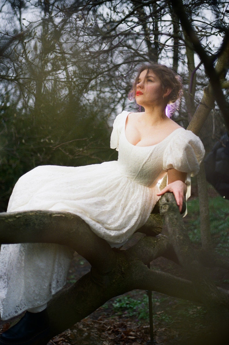 May include: A woman wearing a white lace dress with puffed sleeves sits on a tree branch in a forest setting.