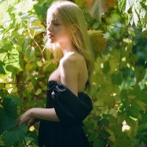May include: A young woman with long blonde hair wearing a black off-the-shoulder dress stands in a lush green garden.