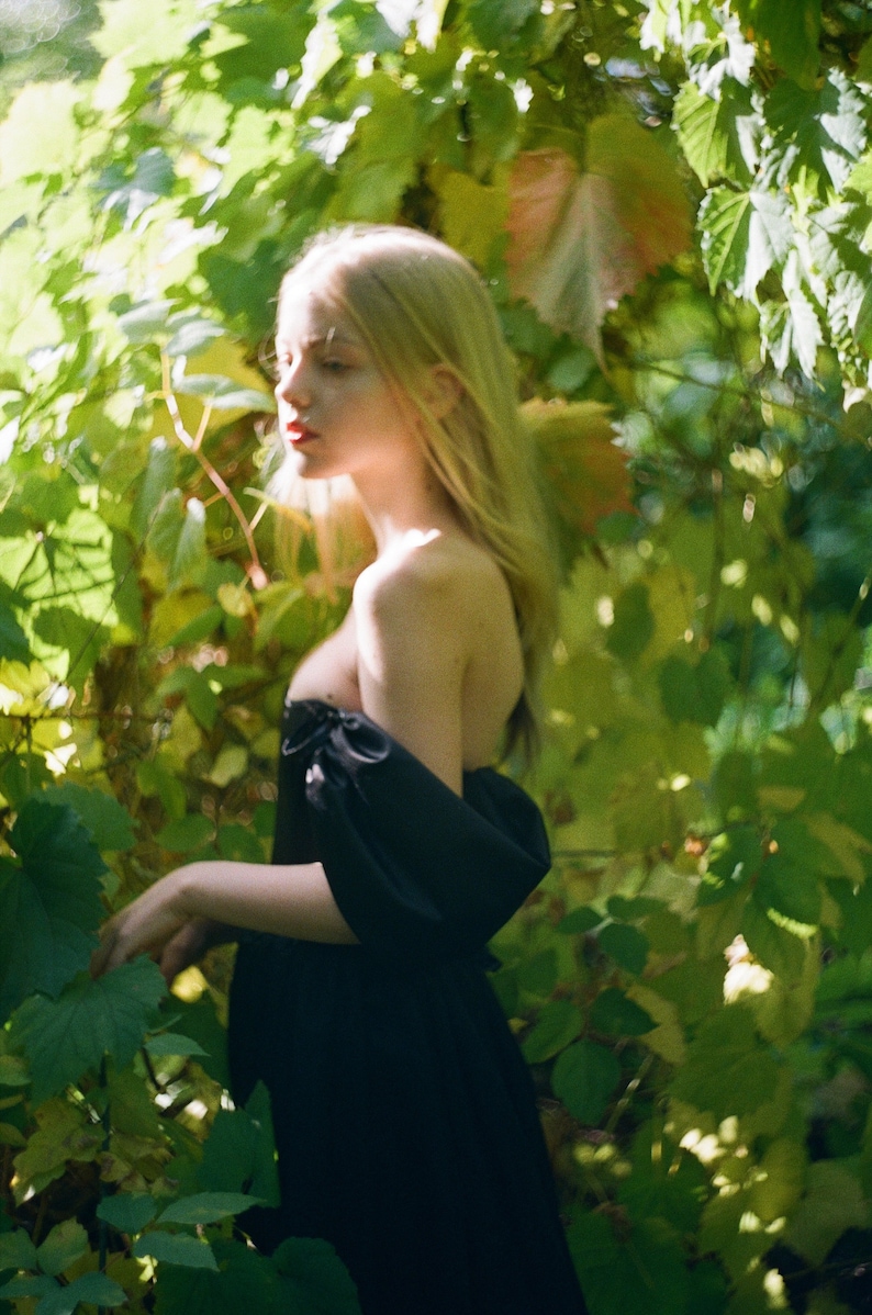 May include: A young woman with long blonde hair wearing a black off-the-shoulder dress stands in a lush green garden.