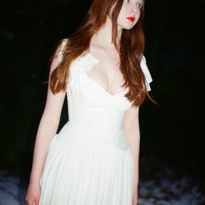 May include: A woman in a white dress with a fitted bodice and flowing skirt stands outdoors. She has long auburn hair and red lipstick. The dress has a sweetheart neckline and ruffled sleeves. The background is dark with a hint of snow.